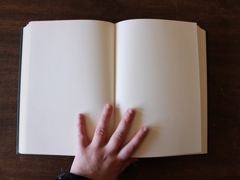 K&ouml;nnte beinhalten: Ein offenes, leeres Buch mit einer Hand, die auf den aufgeschlagenen Seiten ruht. Das Buch hat einen dunkelgr&uuml;nen Einband und wei&szlig;e Seiten.