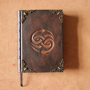 Puede incluir: Un diario encuadernado en cuero marrón oscuro con un diseño de nudo circular en relieve en la portada. El libro incluye protectores de esquina de metal de color latón con estilo antiguo y un marcapáginas de cinta marrón oscuro en el borde inferior.