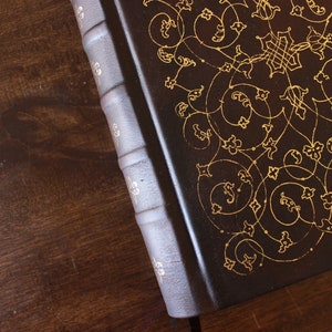K&ouml;nnte beinhalten: Ein dunkelbraunes, ledergebundenes Buch mit goldenen, filigranen Blumenmustern auf dem Einband.
