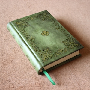 May include: A green hardback book featuring a dark embossed floral border and a central flower motif on the front cover. The book includes a matching green ribbon bookmark extending from the bottom edge, with white pages visible along the side.