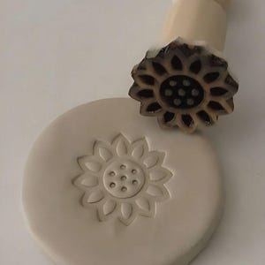 May include: A wooden sunflower stamp with a carved design, next to a clay impression of the same flower. The stamp has a light wood handle and a dark brown flower head. The clay is a light beige color.