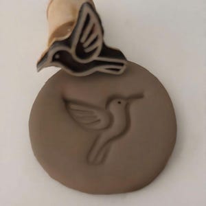 May include: A wooden stamp with a hummingbird design. The stamp has a brown bird image on a round, brown surface. The stamp handle is light brown. The bird is depicted in flight, with its wings spread. The stamp creates an impression of a hummingbird.