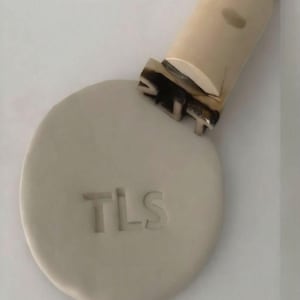 May include: A ceramic stamp with the letters "TLS" imprinted on a round, light gray clay piece. The stamp handle is off-white with a dark brown and silver stamp head.