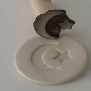 Puede incluir: Un sello de madera clara con un diseño de luna creciente y estrella. El sello se presiona en una pieza redonda de arcilla clara, creando una impresión en relieve de los mismos símbolos celestiales. El mango es de color beige claro.