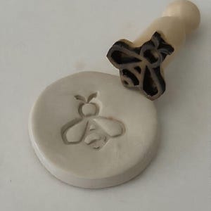 May include: A light beige bee-shaped stamp with a wooden handle sits beside a circular clay impression of a bee. The stamp's design is visible on the clay, showing the bee's outline. The stamp is a craft tool.