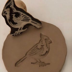May include: A brown bird stamp with a detailed cardinal design, next to an impression of the same bird on a tan clay surface. The stamp has a wooden handle. The bird's features are clearly defined.