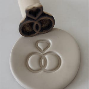 May include: A wooden stamp with a heart and two interlocking rings design, next to a circular clay impression of the same design. The stamp has a wooden handle and a dark brown imprint surface. The clay impression is a light beige color.