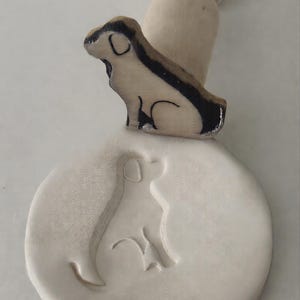 May include: A wooden dog-shaped stamp with a black and brown design, sitting on top of a circular impression of the same dog. A wooden handle is behind the stamp. The stamp creates a dog silhouette.