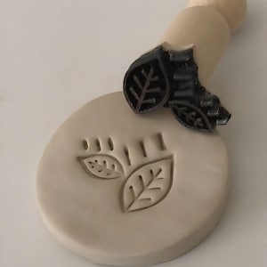 May include: A wooden leaf stamp with a natural wood handle is pressed into a light beige clay disc, creating a leaf imprint. The stamp has a black leaf design. The clay disc has two leaf imprints and three vertical lines.