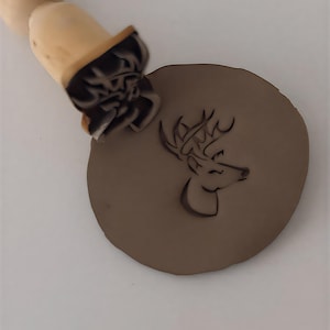 May include: A wooden stamp with a deer head design is pressed into a round piece of brown clay. The stamp has a wooden handle and a detailed carving of a deer's profile. The clay is a solid, earthy color.
