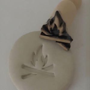 Può includere: Un timbro in ceramica beige chiaro con un disegno di falò. Il timbro ha una base circolare con un'immagine di falò in rilievo. Un pezzo separato con un disegno di fiamma nera e marrone è posizionato sopra la base.