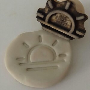 May include: A light-colored, sun-shaped stamp with a matching wooden handle. The stamp's design features a sun with rays and a horizontal line. The wooden handle has a dark brown, burnt appearance.