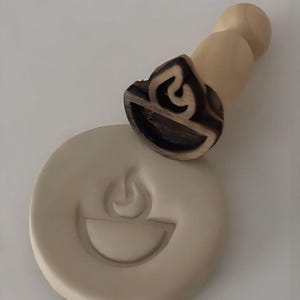 Puede incluir: Un sello de madera con un diseño de llama, junto a una impresión de arcilla de la misma imagen. El sello tiene un mango de madera clara y un diseño de llama oscuro tallado. La impresión de arcilla es de color beige claro.