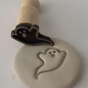 May include: A wooden stamp with a black ghost design is pressed into a round, light-colored clay piece. The stamp has a wooden handle and a detailed ghost shape. The impression of the ghost is visible in the clay.
