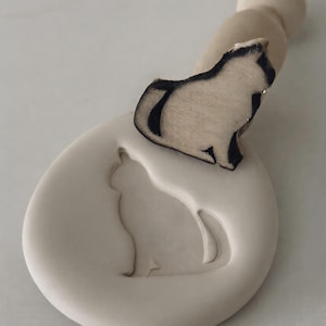 Op de afbeelding: Een houten katvormige stempel met een handvat, met een zwart-wit ontwerp. De stempel staat op een ronde, crèmekleurige basis met een katten silhouet. Ideaal voor knutselen en het toevoegen van een unieke touch.