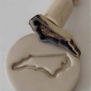 May include: A light-colored wooden cookie stamp featuring the outline of North Carolina. The stamp has a round base and a handle. The top of the stamp has a dark, burnt-looking design.
