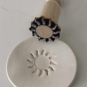 Puede incluir: Un sello de madera con diseño de sol, con mango redondo y patrón de sol detallado. El sello se coloca junto a una pieza redonda de arcilla blanquecina con una impresión de sol. Ideal para manualidades y proyectos decorativos.