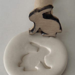 Op de afbeelding: Een houten konijnenstempel met een uitgesneden konijnenontwerp, gebruikt om een konijnenvorm in een ronde, crèmekleurige kleischijf te drukken. De stempel heeft een houten handvat en een konijnensilhouet met zwarte details.