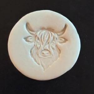 May include: A round, off-white ceramic stamp featuring a detailed relief of a Highland cow's head. The cow has long, shaggy fur and curved horns. The stamp is circular and appears to be handmade.