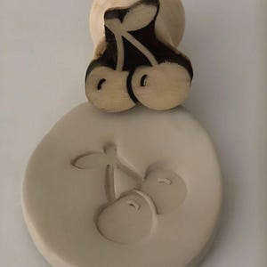 Puede incluir: Un sello de madera con forma de cereza y un mango redondo, junto a una impresión circular del mismo diseño de cereza. El sello presenta un diseño detallado de cereza con detalles en marrón oscuro, y la impresión es de color beige claro.