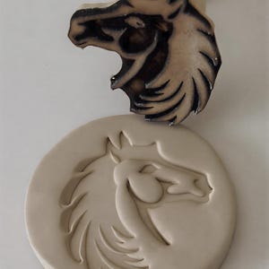 Puede incluir: Un sello de madera con forma de cabeza de caballo con un mango, junto a una impresión circular de arcilla de una cabeza de caballo. El sello tiene un diseño tallado detallado con acentos oscuros, mientras que la impresión es de un color más claro y neutro.