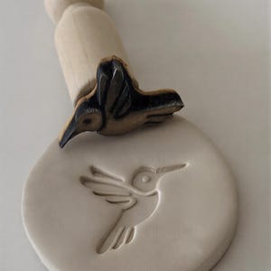 May include: A wooden stamp with a hummingbird design. The stamp features a carved hummingbird on top and an imprint of the same design on the base. The handle is a light wood color, and the hummingbird is brown and black.