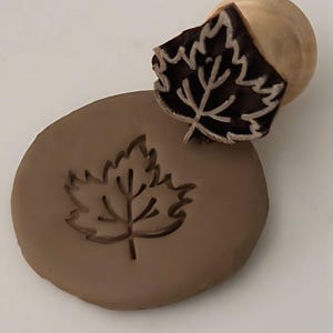 Puede incluir: Un sello de madera con forma de hoja con una impresión de hoja marrón sobre un disco de arcilla marrón. El sello tiene un mango de madera redondeado. El diseño de la hoja está detallado con venas y un borde texturizado.