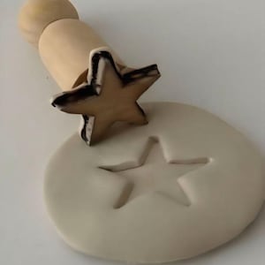 Könnte beinhalten: Ein Holzstempel in Sternform mit einem hellbraunen Griff wird in einen hellbeigen Ton gedrückt, wodurch ein Sternabdruck entsteht. Der Stempel hat eine dunkle Umrandung, und der Ton befindet sich auf einer weißen Oberfläche.