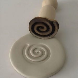 Könnte beinhalten: Ein hellbeige Keramikstempel mit einem Spiralmuster. Der Stempel hat einen Holzgriff und ein dunkelbraunes Spiralmuster auf der Unterseite. Ein passendes hellbeige Keramikstück mit einem Spiralmuster ist ebenfalls vorhanden.