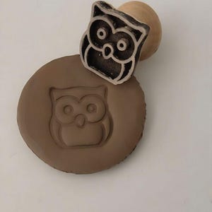 May include: A wooden owl stamp with a round handle is pressed into a brown clay circle, creating an owl imprint. The stamp is black with white details. The clay is a light brown color.
