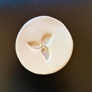 May include: A circular white ceramic disc featuring a debossed triskelion-style leaf motif in the center. The surface of the disc is smooth and matte, with the three-pointed floral design pressed into the material to create depth and shadow.