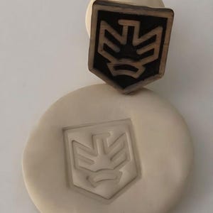 May include: A wooden stamp with a shield-shaped design, featuring a geometric emblem, is pressed into a circle of clay. The stamp is dark brown with a light brown emblem, and the impression in the clay is a lighter shade.