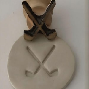 May include: A beige golf-themed decorative item. The item features a round base with an embossed image of crossed golf clubs. Above the base, two miniature golf clubs are crossed. The golf clubs are brown with black accents.