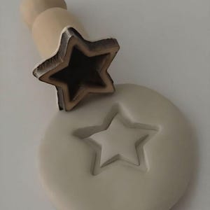 May include: A wooden star-shaped stamp with a handle, next to a round, light-colored clay piece with a star imprint. The stamp has a dark brown outline and a hollow center. The clay piece has a raised star design.