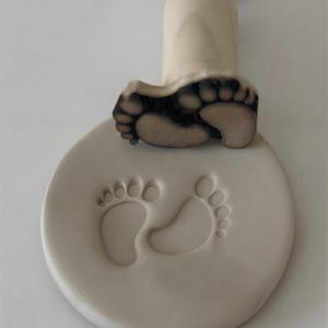 May include: A light beige baby footprint stamp with a wooden handle. The stamp has a detailed impression of a baby's foot, and the base has an imprint of two baby footprints. The stamp is designed for creating keepsakes.