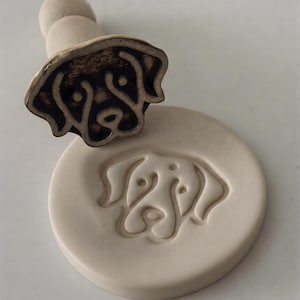 May include: A ceramic dog stamp with a wooden handle, next to a matching impression. The stamp features a detailed outline of a dog's face. The stamp and impression are in shades of brown and cream.