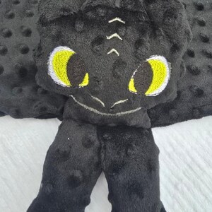 May include: A black plush dragon-themed baby blanket with yellow eyes and white accents. The blanket features a dragon head and two feet, all made of soft, textured fabric. The background is a gray dotted fabric.