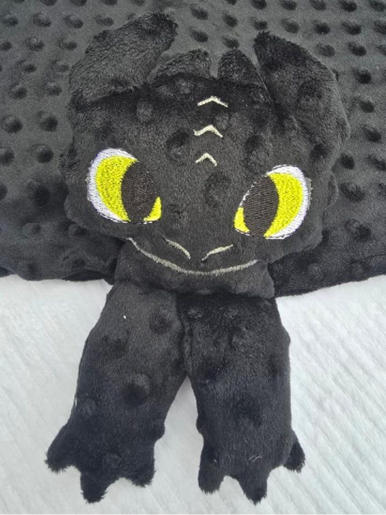 May include: A black plush dragon-themed baby blanket with yellow eyes and white accents. The blanket features a dragon head and two feet, all made of soft, textured fabric. The background is a gray dotted fabric.