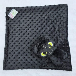 May include: A black baby blanket with a textured, dotted surface. A plush toy in the shape of a dragon with yellow eyes and white accents is attached to the blanket. A small tag is in the upper left corner.