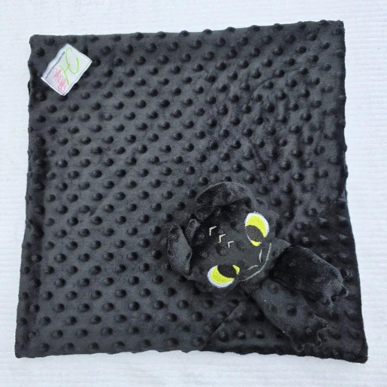 May include: A black baby blanket with a textured, dotted surface. A plush toy in the shape of a dragon with yellow eyes and white accents is attached to the blanket. A small tag is in the upper left corner.