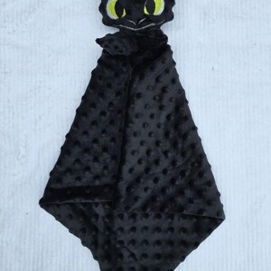 May include: A black plush baby security blanket featuring a dragon head with yellow eyes. The blanket has a textured, dotted pattern and a soft, cuddly appearance. The dragon head is embroidered with details.
