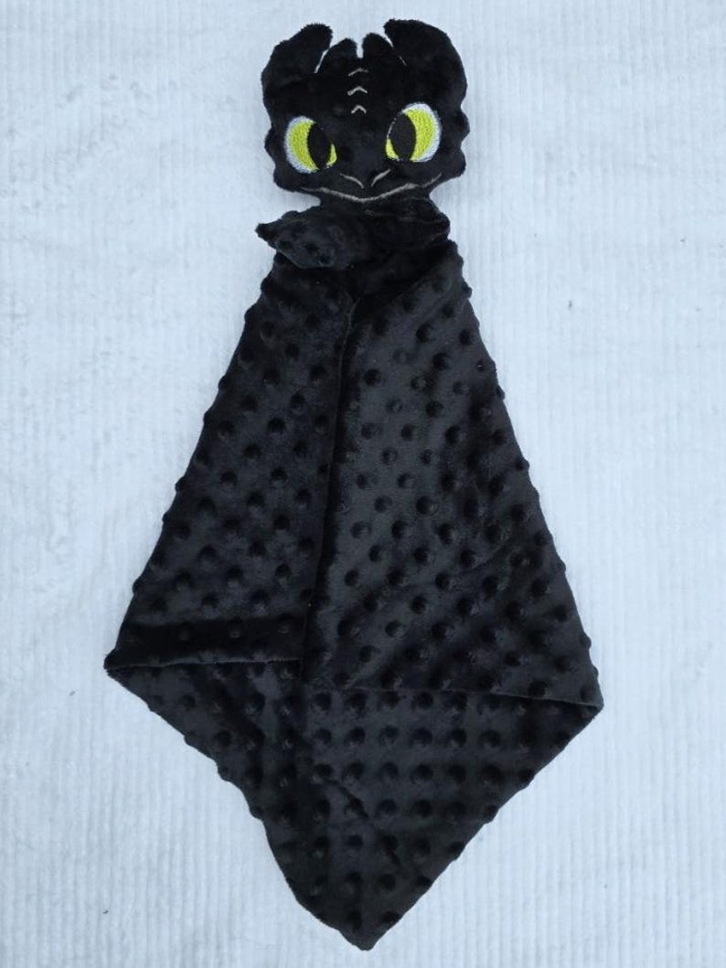 May include: A black plush baby security blanket featuring a dragon head with yellow eyes. The blanket has a textured, dotted pattern and a soft, cuddly appearance. The dragon head is embroidered with details.