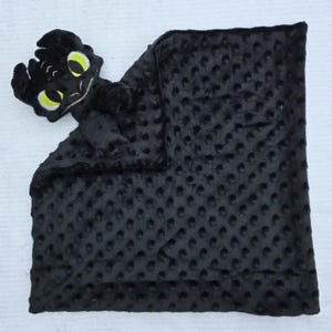 May include: A black baby blanket with a plush, textured surface and a dragon head attached. The dragon head has yellow eyes and is made of soft material. The blanket is approximately 20 inches square.