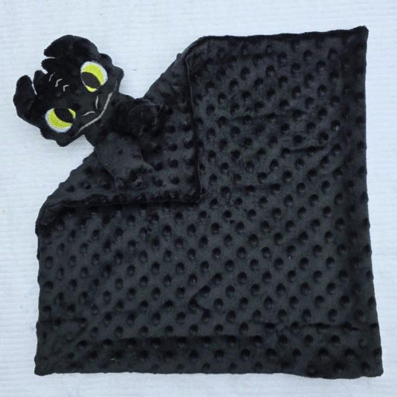 May include: A black baby blanket with a plush, textured surface and a dragon head attached. The dragon head has yellow eyes and is made of soft material. The blanket is approximately 20 inches square.