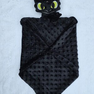 May include: A black baby security blanket featuring a plush dragon head with yellow eyes. The blanket is made of a soft, textured fabric with a dotted pattern. The dragon's head is detailed with embroidered features.