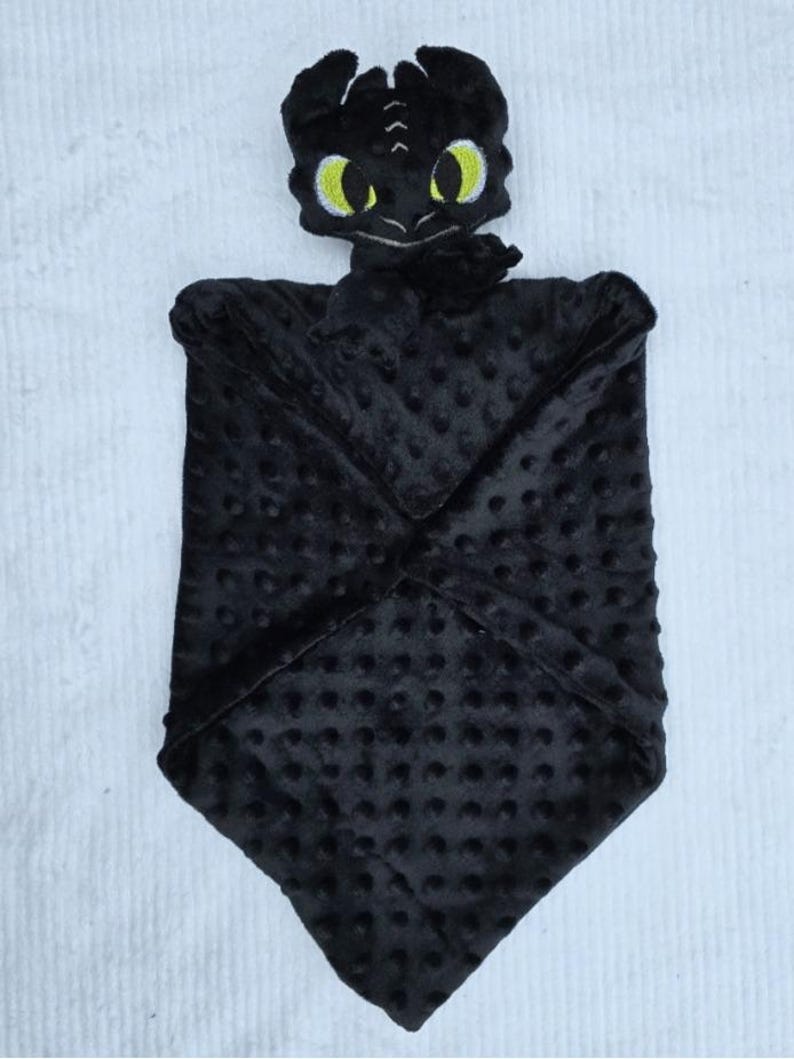 May include: A black baby security blanket featuring a plush dragon head with yellow eyes. The blanket is made of a soft, textured fabric with a dotted pattern. The dragon's head is detailed with embroidered features.