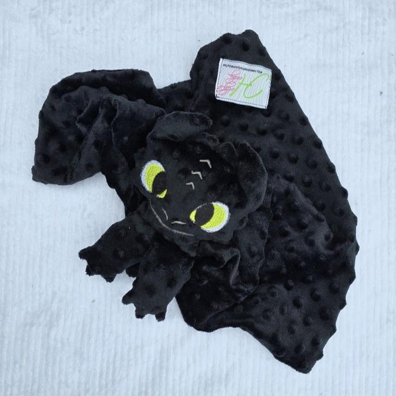 May include: A black baby blanket with a plush, textured surface and a character face. The character has large yellow eyes and is embroidered with white thread. A small tag is sewn on the blanket.