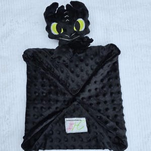 May include: A black plush baby blanket featuring a dragon head with yellow eyes. The blanket has a textured, dotted surface and a square shape. A small label is sewn on the blanket.