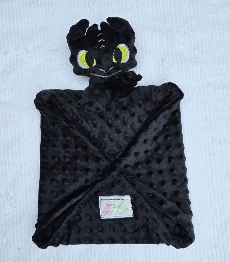 May include: A black plush baby blanket featuring a dragon head with yellow eyes. The blanket has a textured, dotted surface and a square shape. A small label is sewn on the blanket.