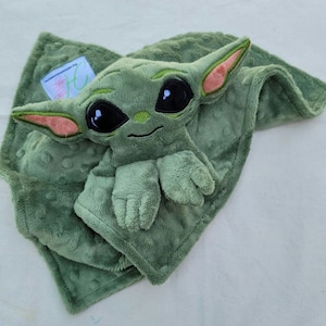 May include: A green plush baby character toy with large black eyes, large ears with pink accents, and small hands and feet, resting on a matching green, textured blanket. The blanket has a small tag with text.